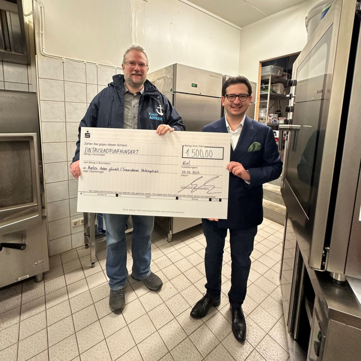 Bastian Mahmoodi überreicht gemeinsam mit einem Vertreter des Kieler Ankers einen symbolischen Spendenscheck über 1.500 Euro in einer Großküche.