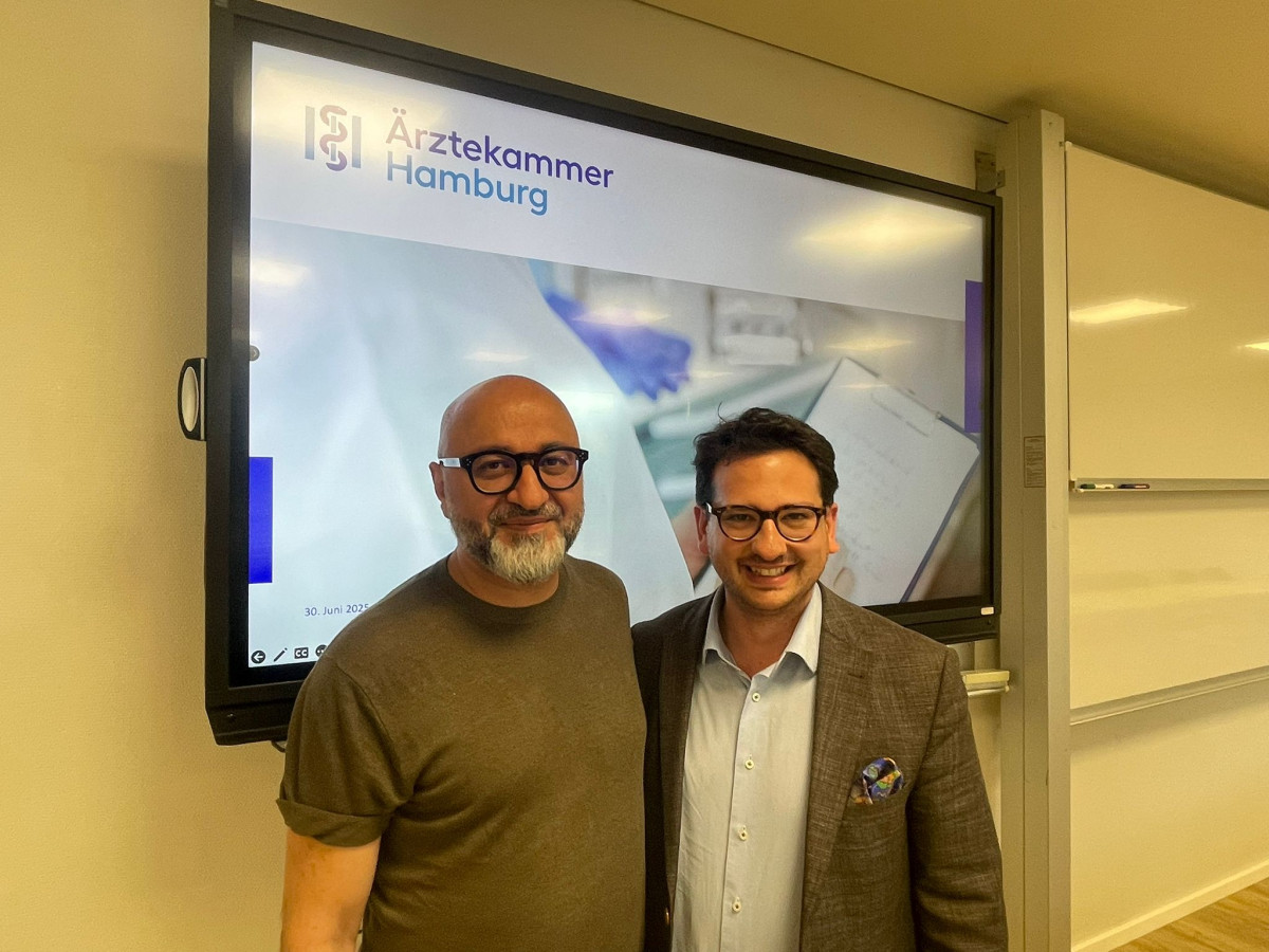 Bastian Mahmoodi und Dr. Pedram Emami stehen lächelnd vor einer Präsentationsfolie der Ärztekammer Hamburg.