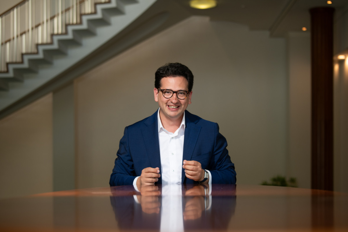 Bastian Mahmoodi sitzt lächelnd an einem Tisch in einem hellen Raum mit Treppe im Hintergrund.