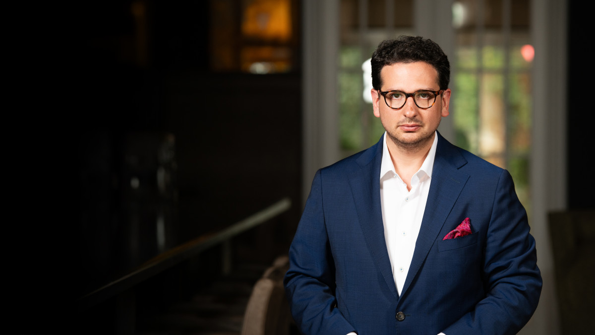 Bastian Mahmoodi steht in einem eleganten Innenraum, trägt einen dunkelblauen Anzug und blickt ruhig in die Kamera.
