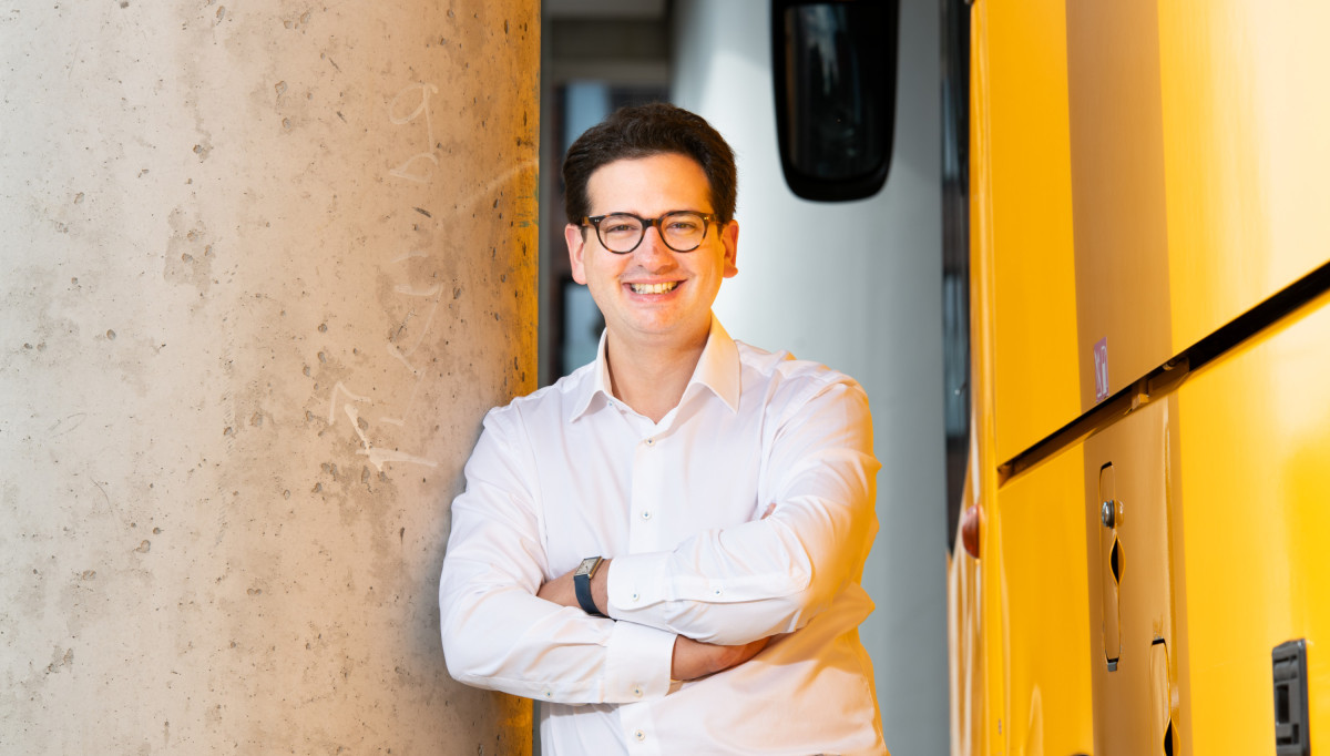 Bastian Mahmoodi lehnt lächelnd mit verschränkten Armen an einer Betonwand neben einem gelben Bus.