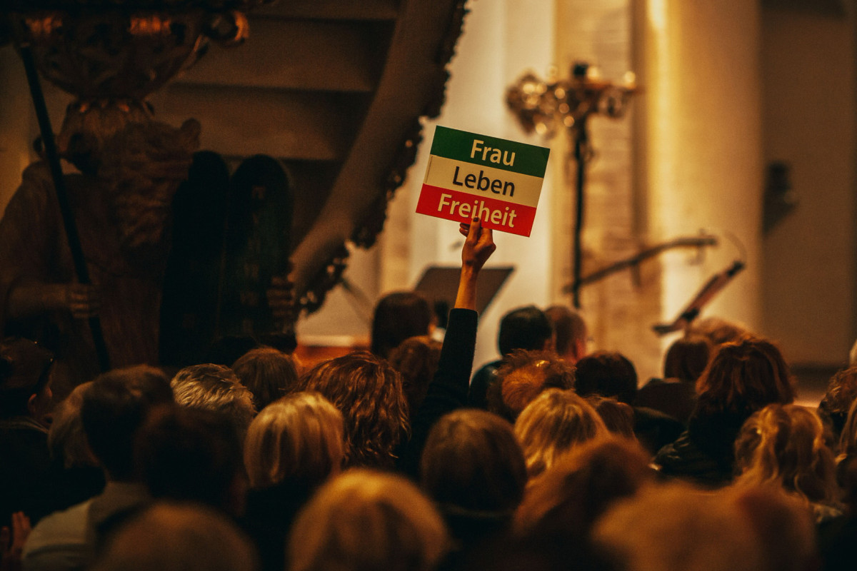 Eine Menschenmenge in einer Kirche. Eine Frau hält ein Schild hoch mit der iranischen Flagge und der Aufschrift „Frau, Leben, Freiheit“.