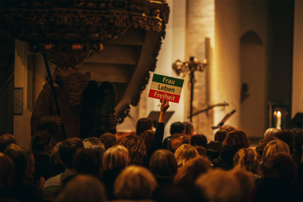 Eine Menschenmenge in einer Kirche. Eine Frau hält ein Schild hoch mit der iranischen Flagge und der Aufschrift „Frau, Leben, Freiheit“.