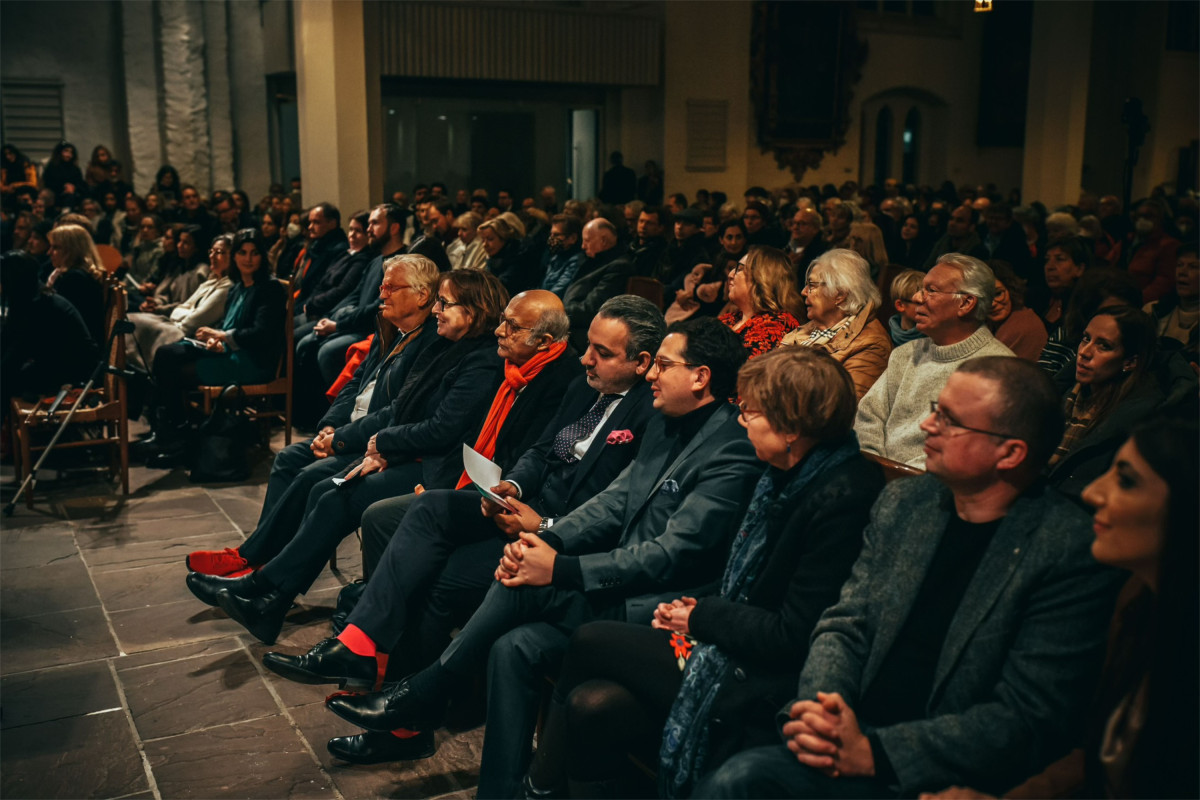 Bastian Mahmoodi sitzt mit weiteren Personen in einem Veranstaltungsraum.