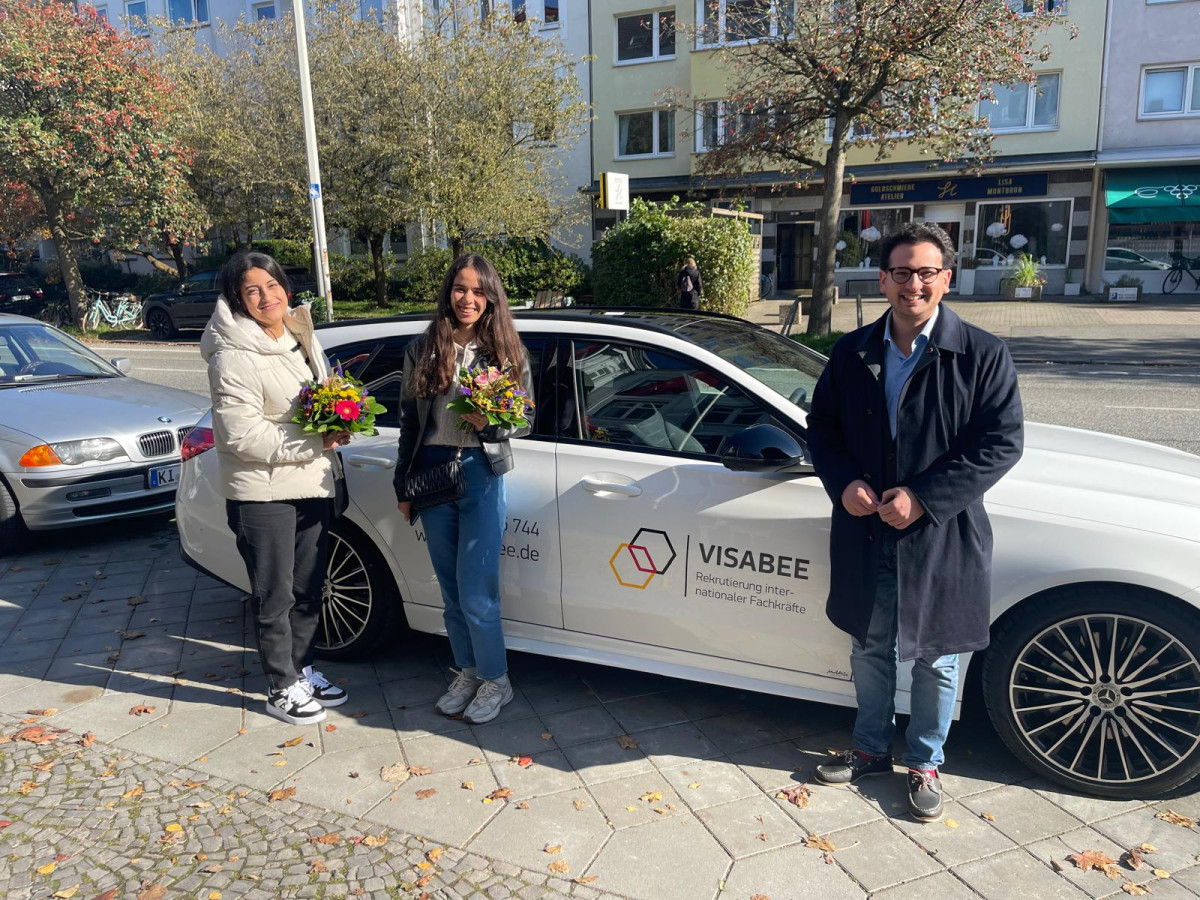Bastian Mahmoodi steht mit zwei Frauen neben einem Auto mit VISABEE-Logo, alle halten Blumensträuße.